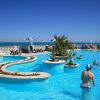 Piscina all'aperto con isole di palme, sedie a sdraio e persone che nuotano.
