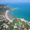 Vista aerea di una spiaggia a forma di mezzaluna, mare azzurro e vegetazione circostante.