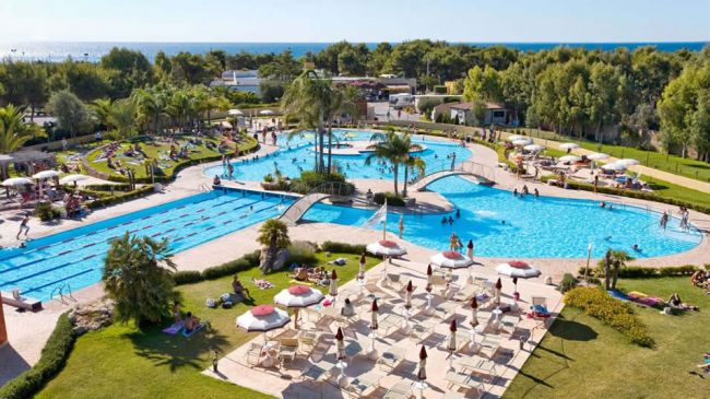 Ampia piscina con palme e ombrelloni, alberi sullo sfondo.