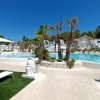 Piscina circondata da palme e piante con cielo blu. Strutture bianche in pietra sullo sfondo.