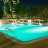 Piscina illuminata di notte, con lettini intorno e alberi sullo sfondo.